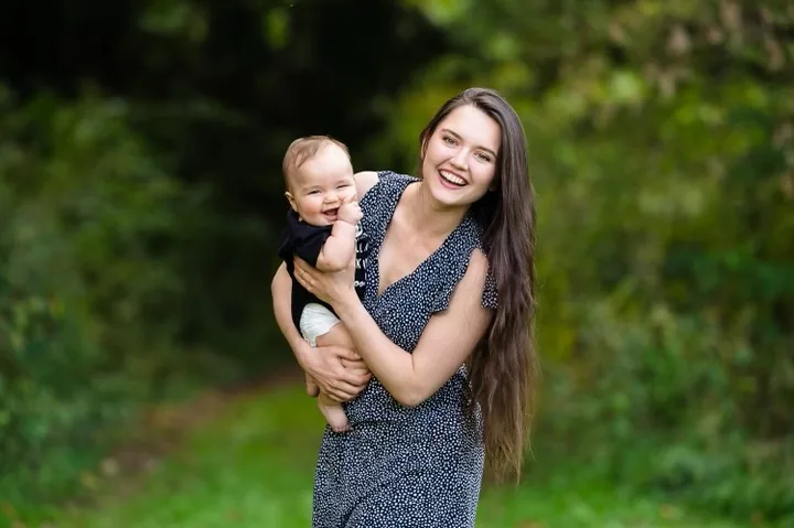A mother smiling outdoors while holding her baby